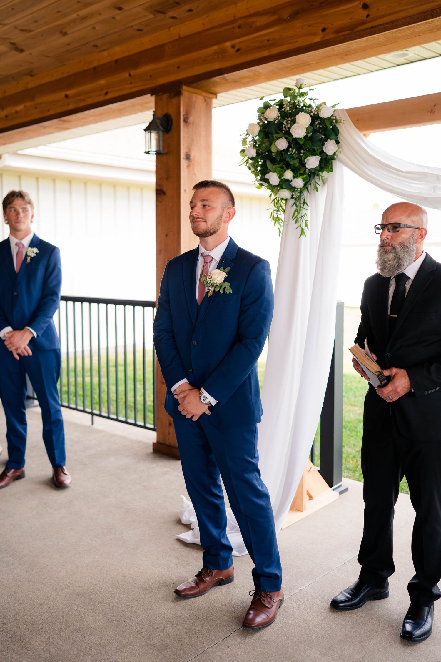 Groom in navy blue suit stands at altar with officiant during outdoor wedding ceremony.