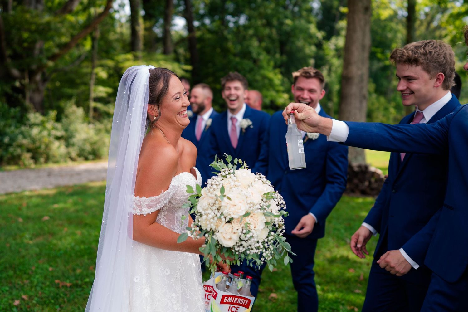 Bride in white gown laughing with groomsmen in navy suits outdoors at wedding celebration.
