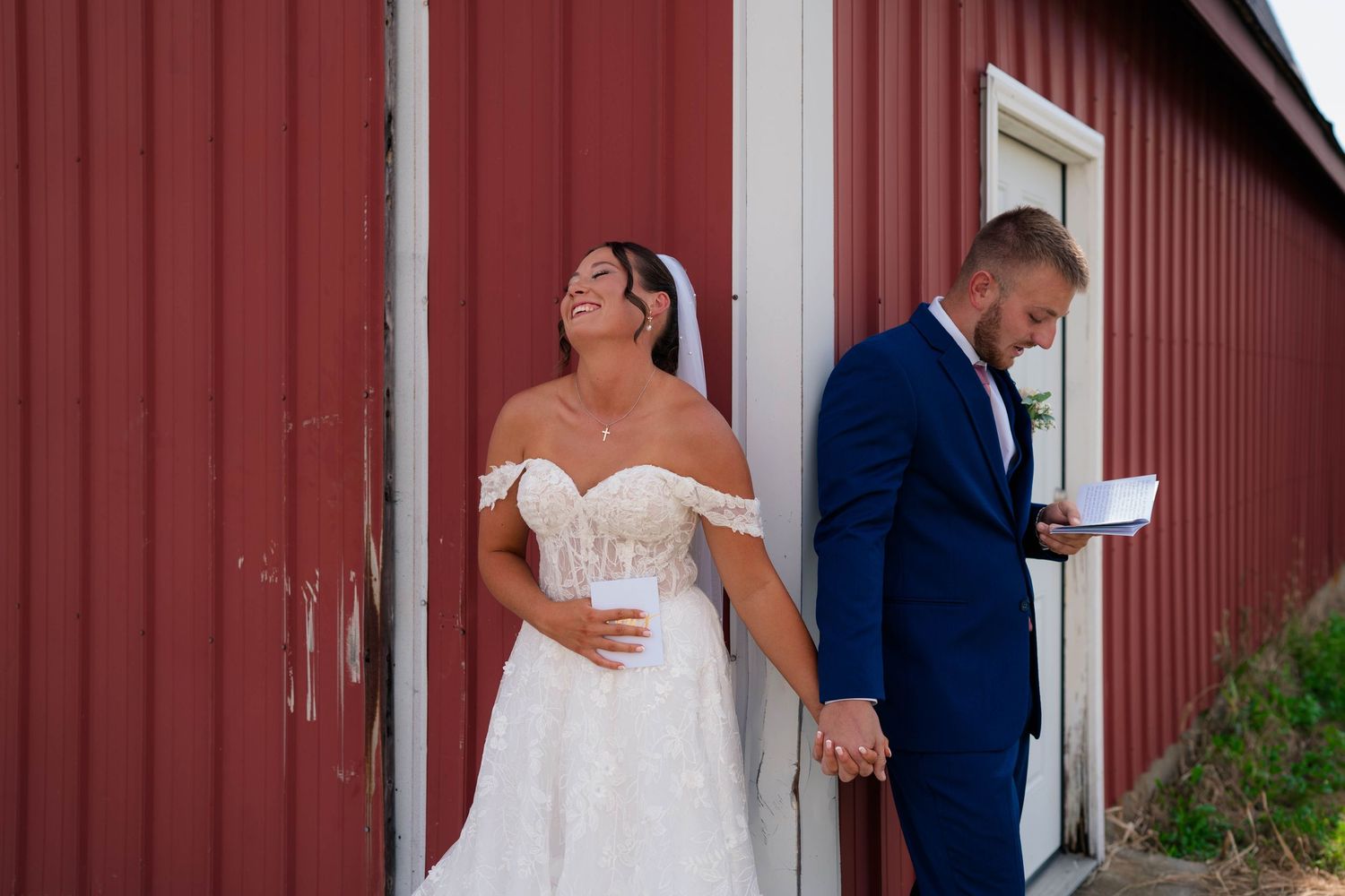 Bride and groom sharing first look moment holding hands against rustic red barn exterior.