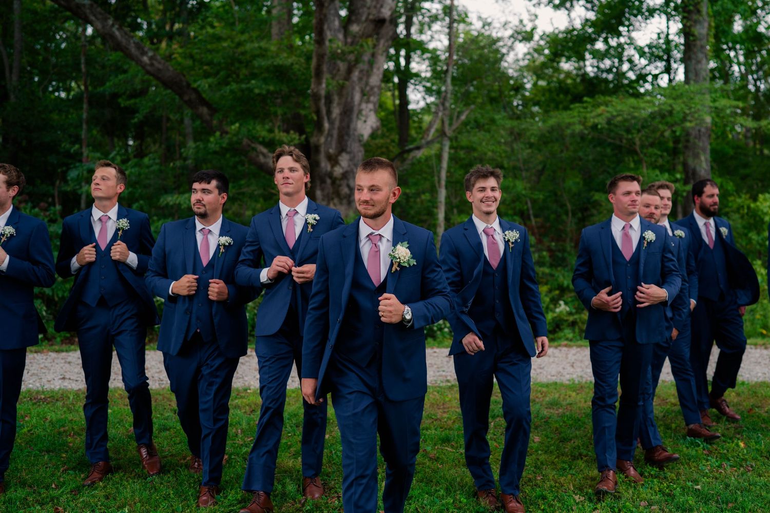 Groom leads groomsmen in navy blue suits with pink ties walking outdoors at a wedding.