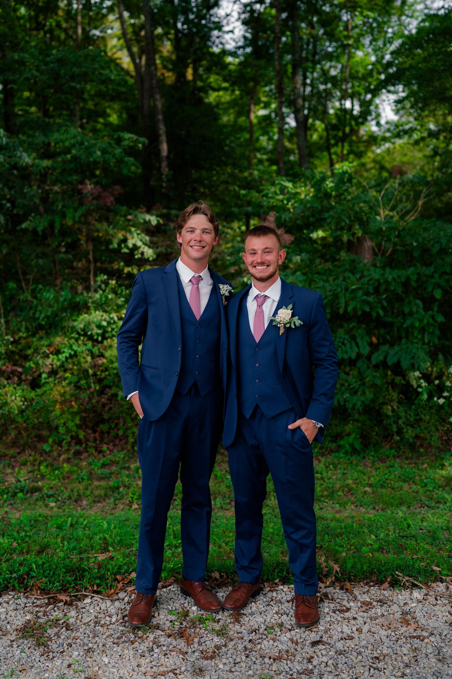 Two groomsmen in matching navy blue suits and pink ties posing outdoors at wedding.