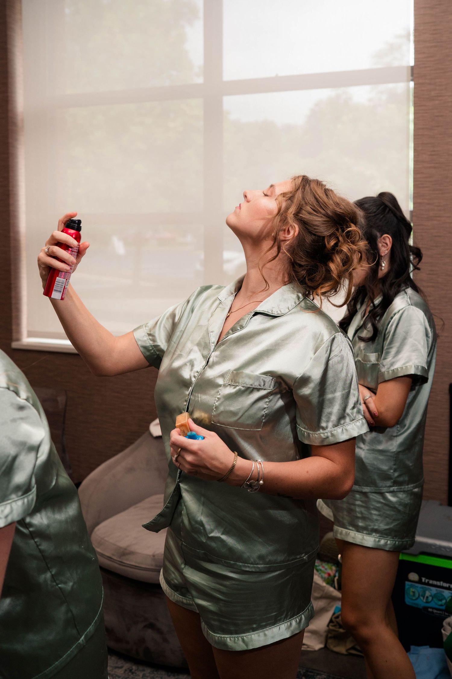 Bridesmaid in sage satin robe applying hairspray while getting ready for a wedding.