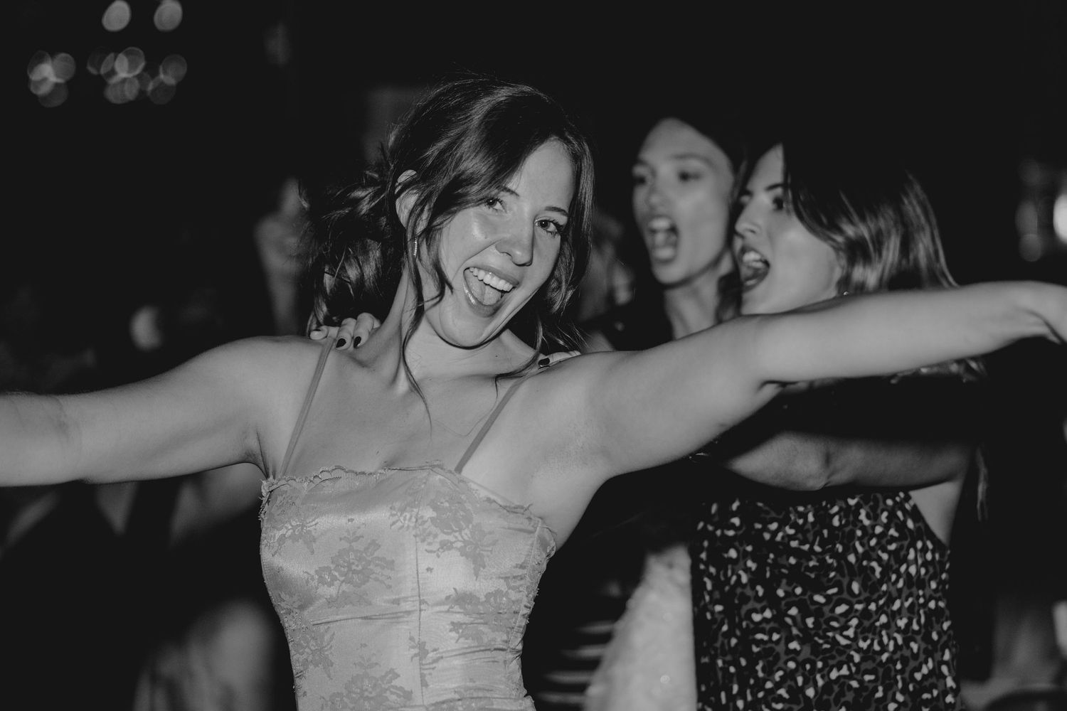 Black and white photo of smiling bride dancing joyfully with friends at wedding reception.