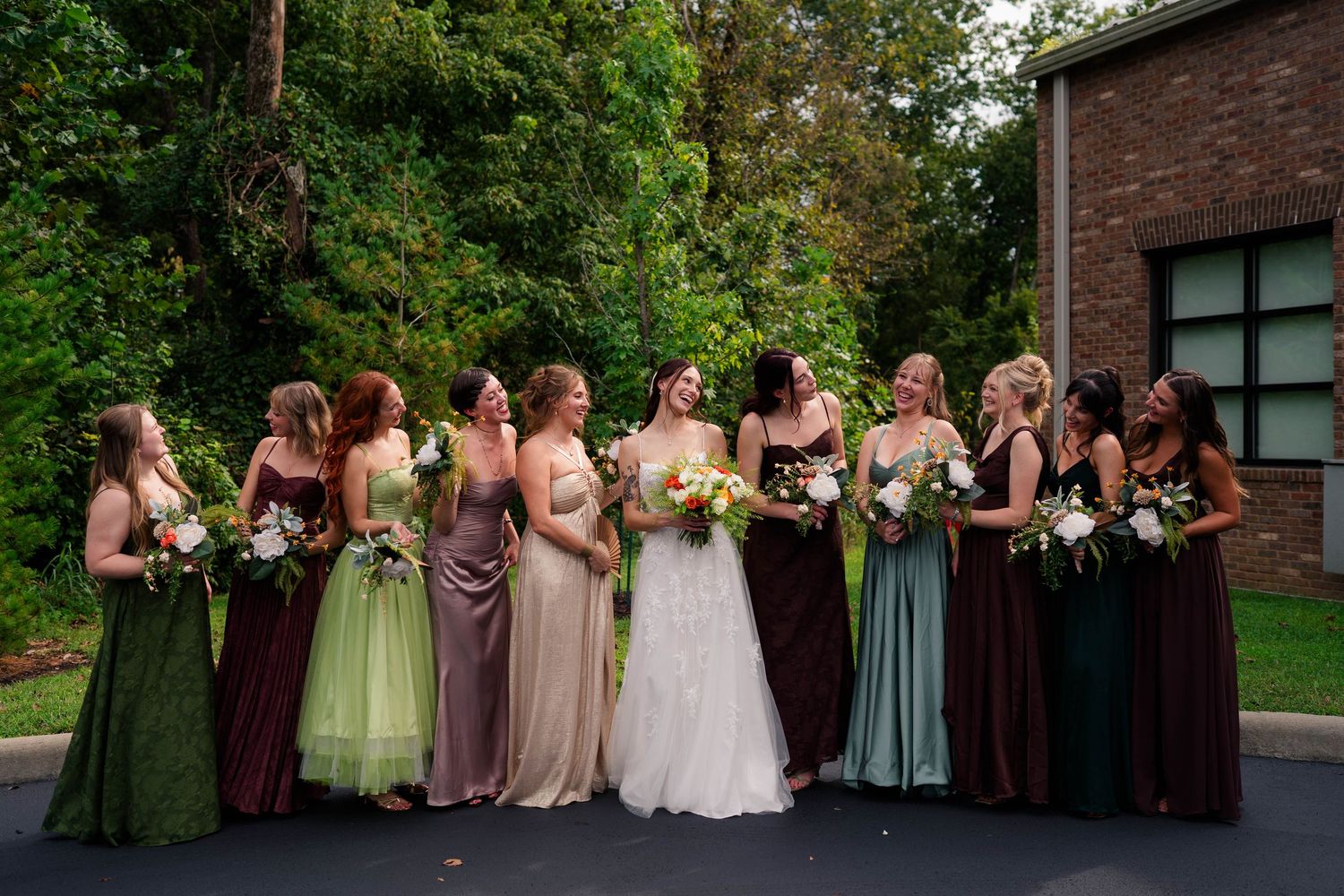 Bride surrounded by bridesmaids in mismatched earth-tone gowns laughing outdoors at wedding celebration.