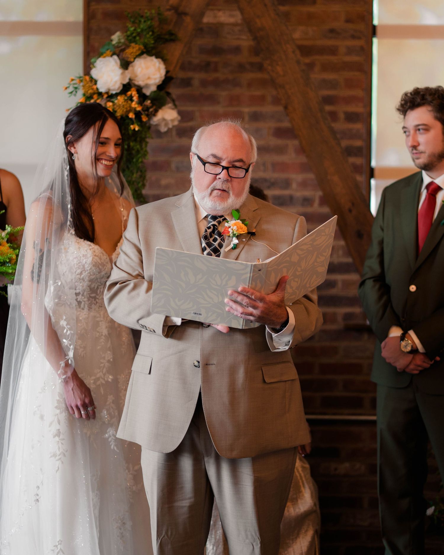 Officiant reads vows as bride smiles during indoor wedding ceremony near floral arch backdrop.