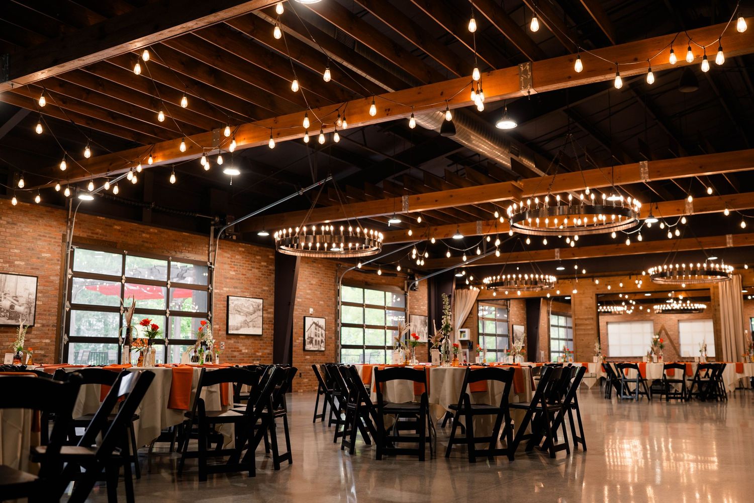 Elegant wedding reception hall with exposed brick, string lights, chandeliers, and black chairs.