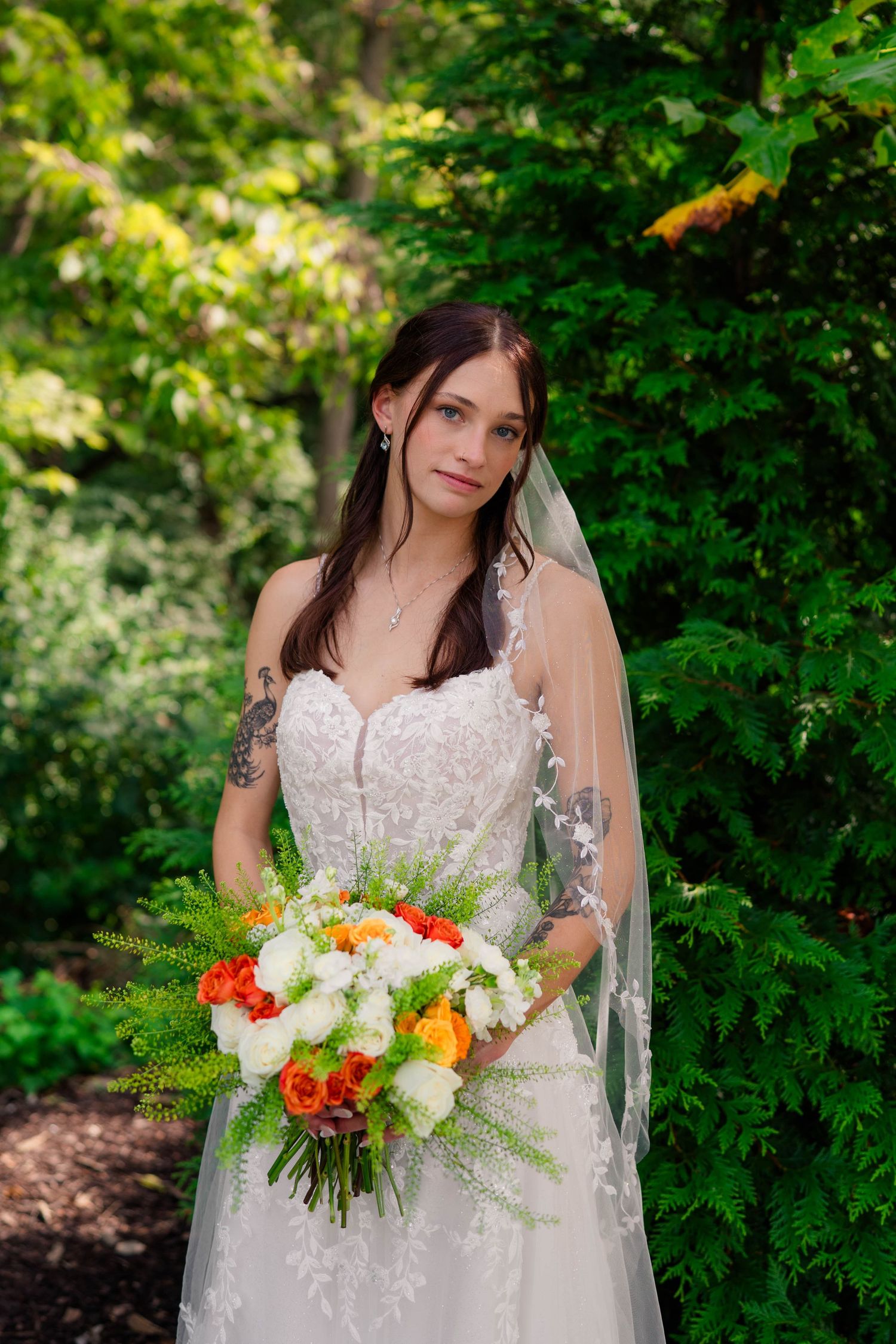 Beautiful bride in lace wedding gown holding colorful orange and white bouquet outdoors.