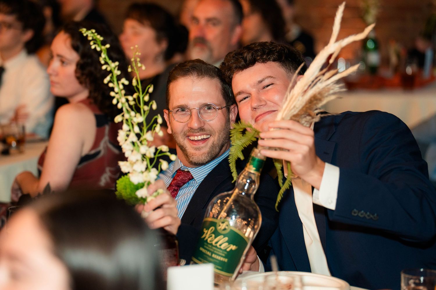Two smiling male wedding guests holding floral centerpiece and whiskey bottle at reception.