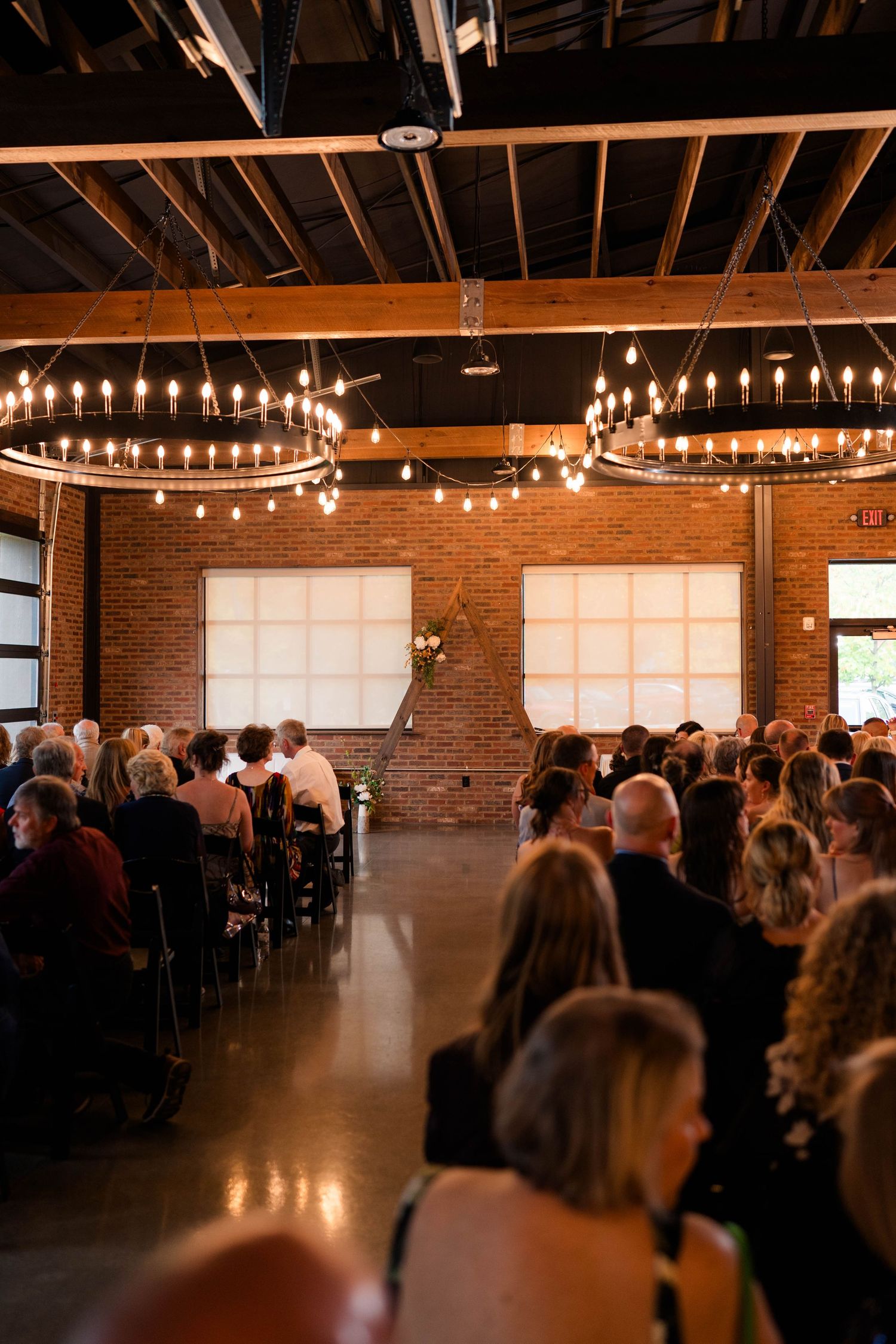 Guests seated in rows at an indoor wedding ceremony in a rustic brick venue with warm Edison bulb chandeliers.