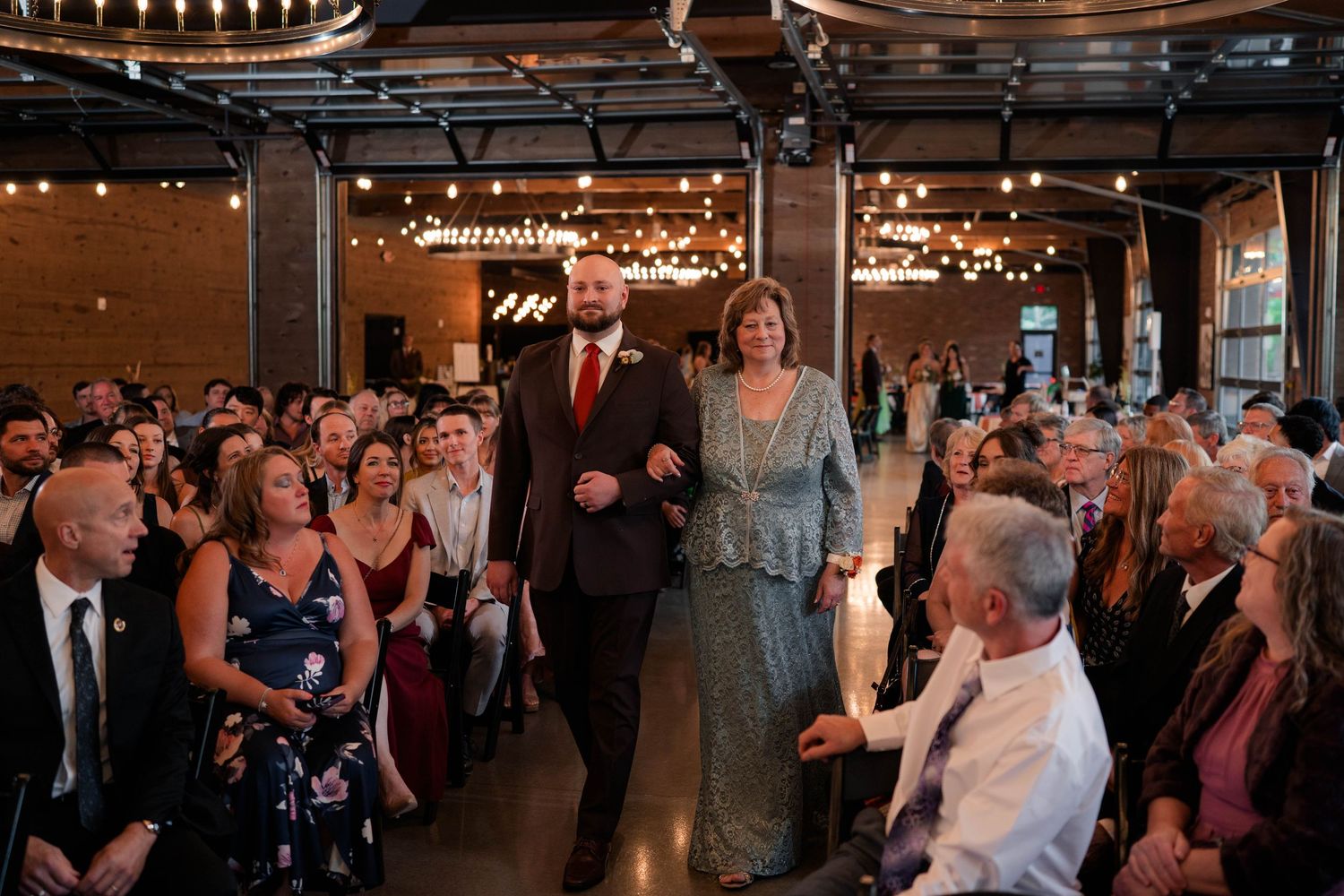 Groom escorts his mother down the aisle at an indoor wedding ceremony with string lights.