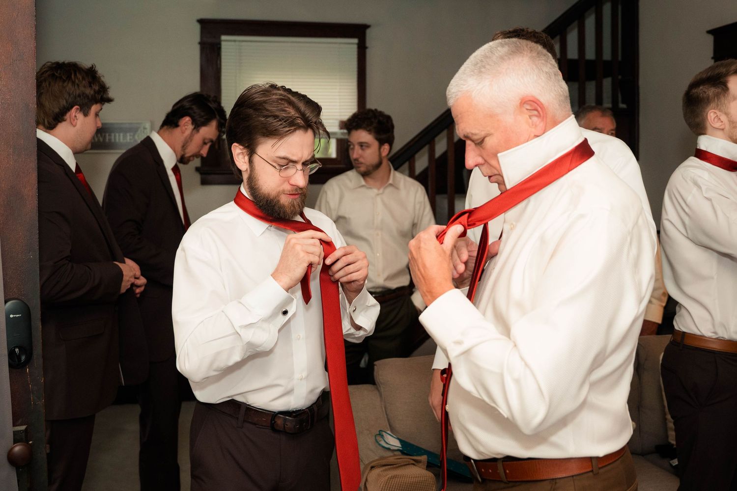 Two men in white dress shirts adjusting red neckties while others prepare in the background.