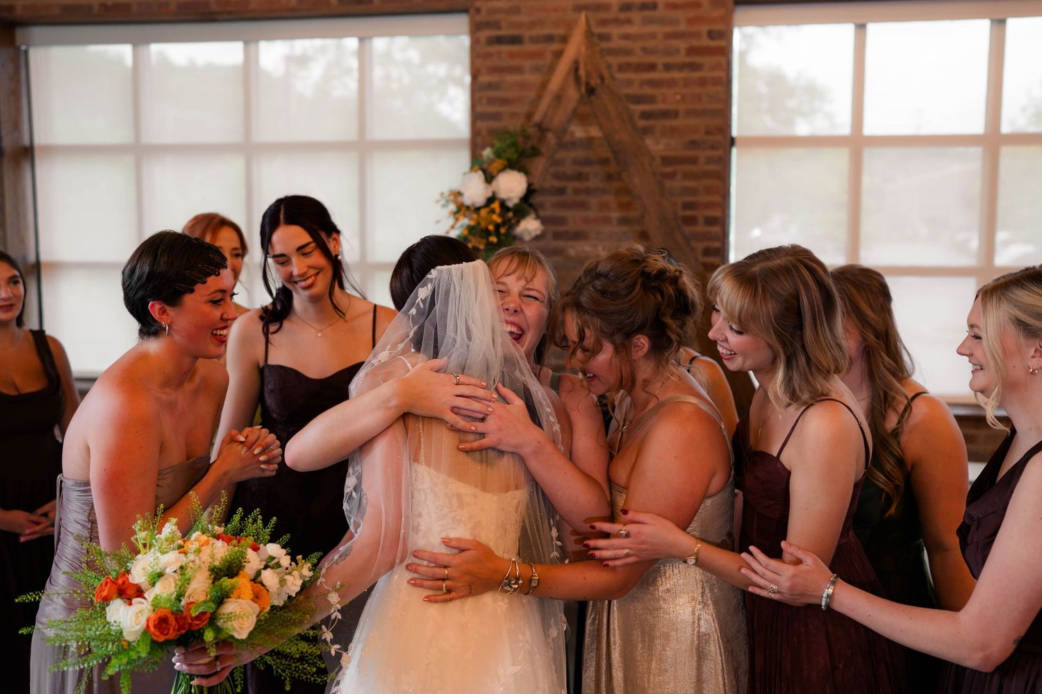 Bride in white gown and veil embraced by bridesmaids in mauve dresses at wedding venue.