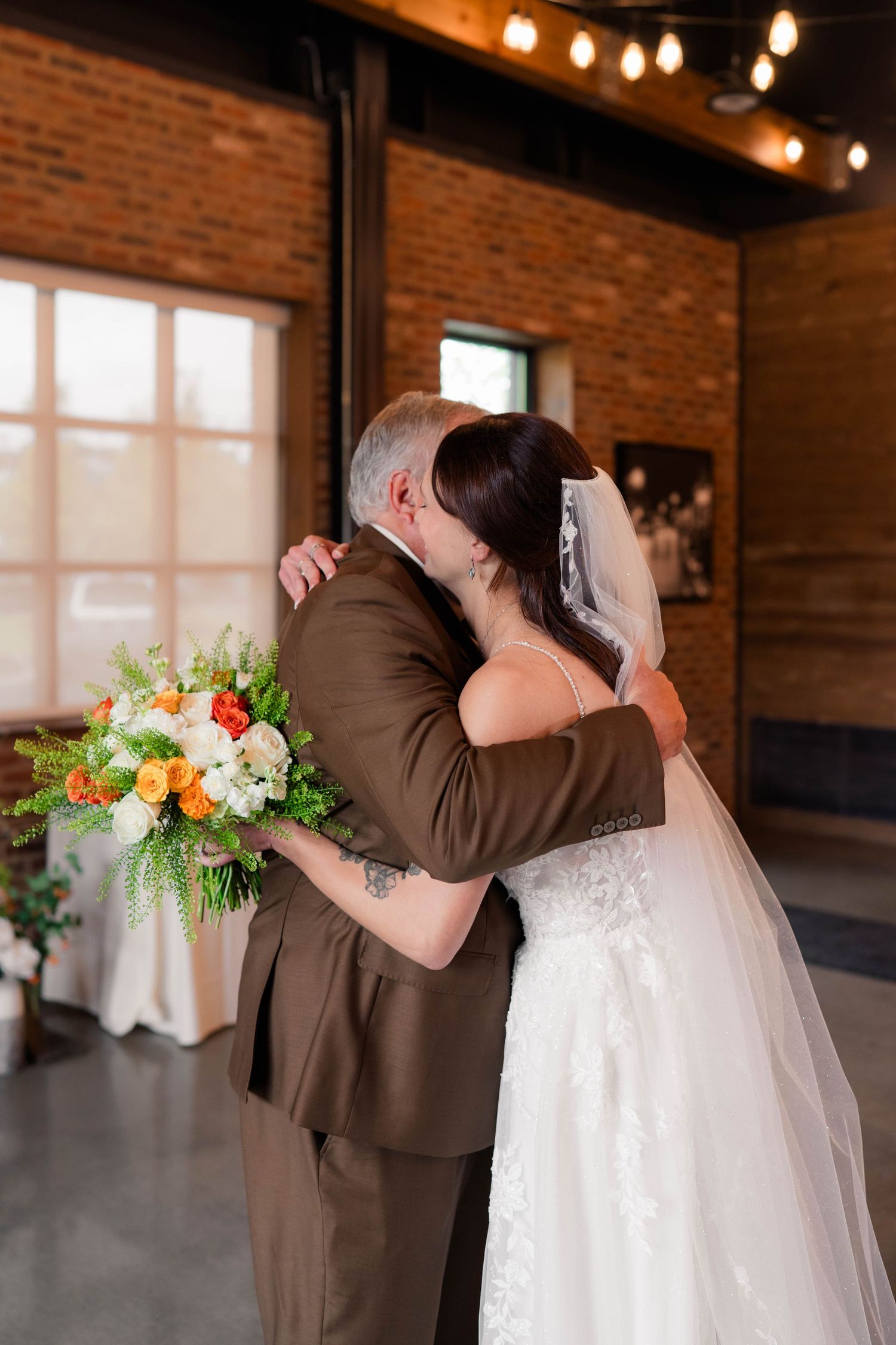 Bride in lace gown embracing her father during an emotional first look in a brick wedding venue.