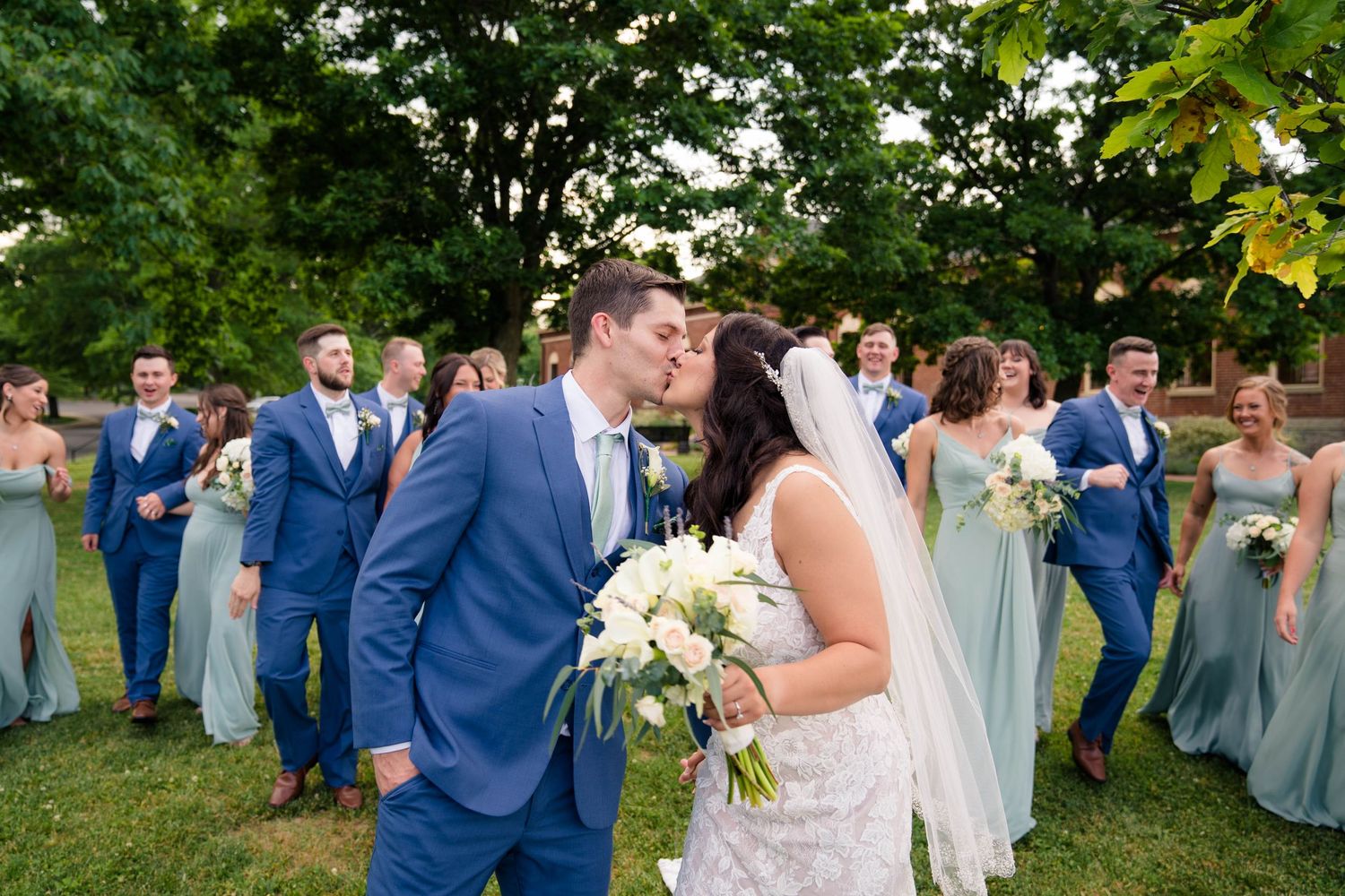 Bride and groom share a kiss surrounded by wedding party in blue suits and sage green bridesmaid dresses outdoors.