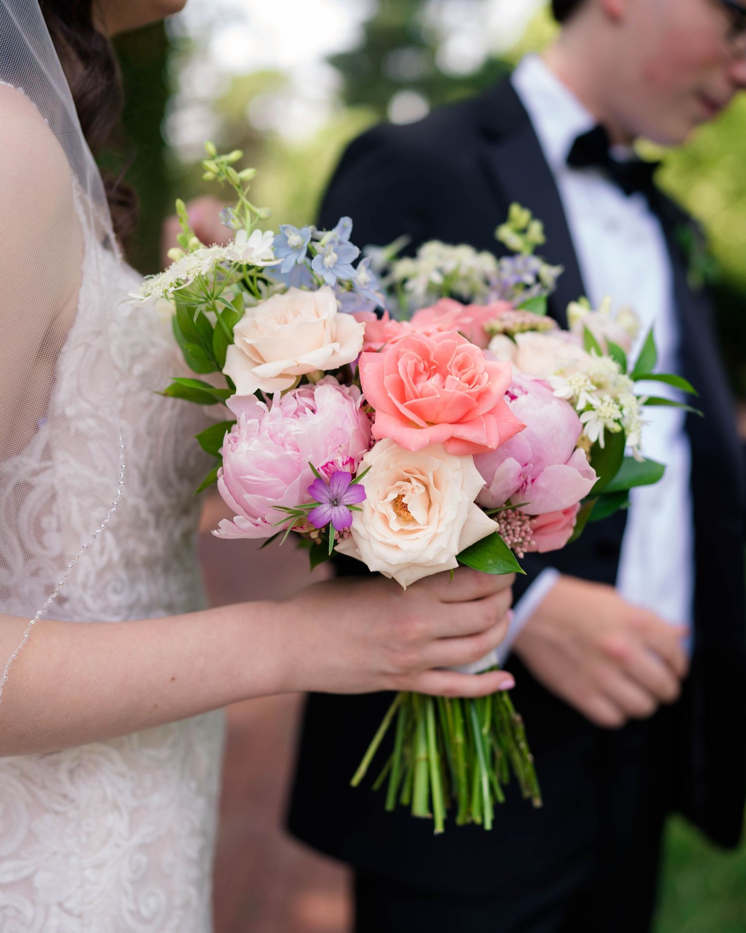 Bride holds a lush pink peony and rose bouquet with blue accents beside groom in black tuxedo.