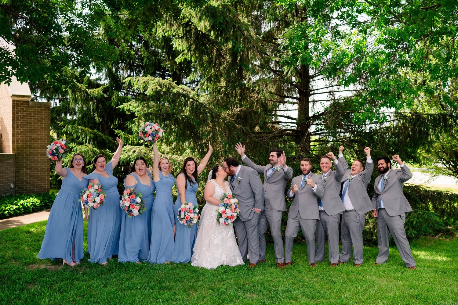 Wedding party in blue dresses and gray suits celebrate joyfully on green lawn under trees.
