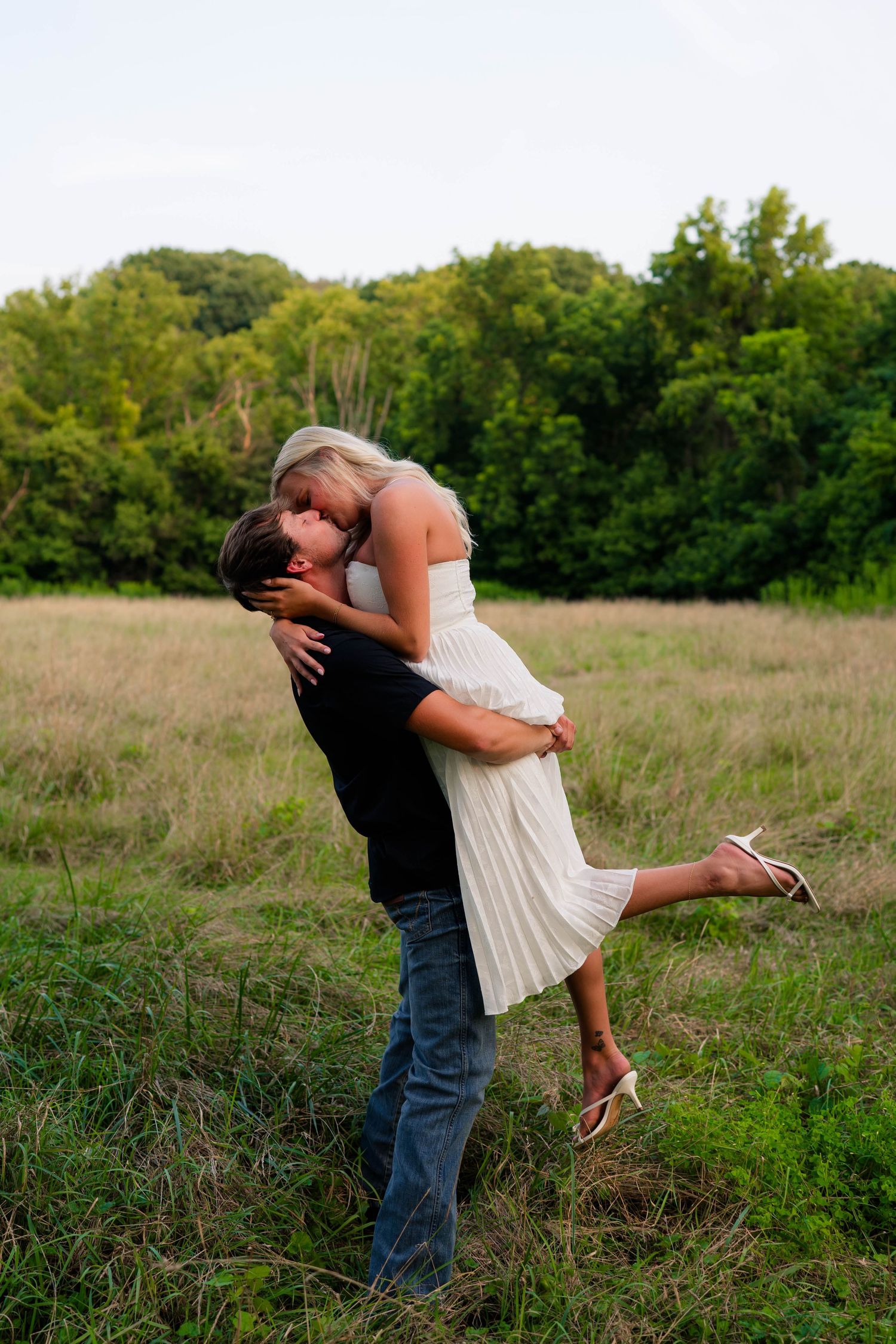 Romantic couple kissing in a grassy field, man lifting woman in white dress outdoors.