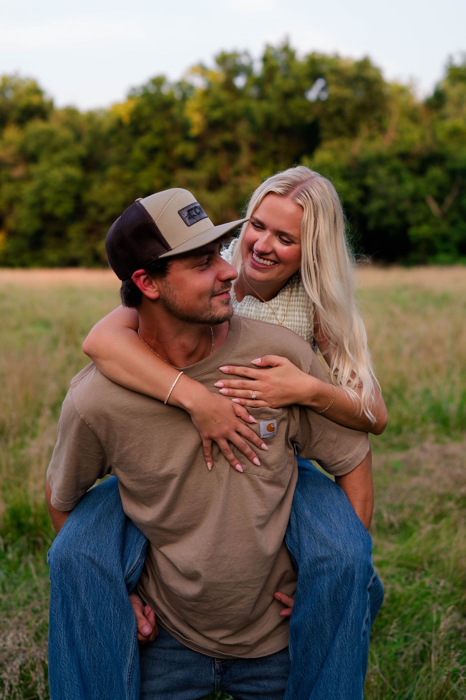 A young couple sharing a piggyback ride in a golden field, smiling joyfully with lush green trees in the background.