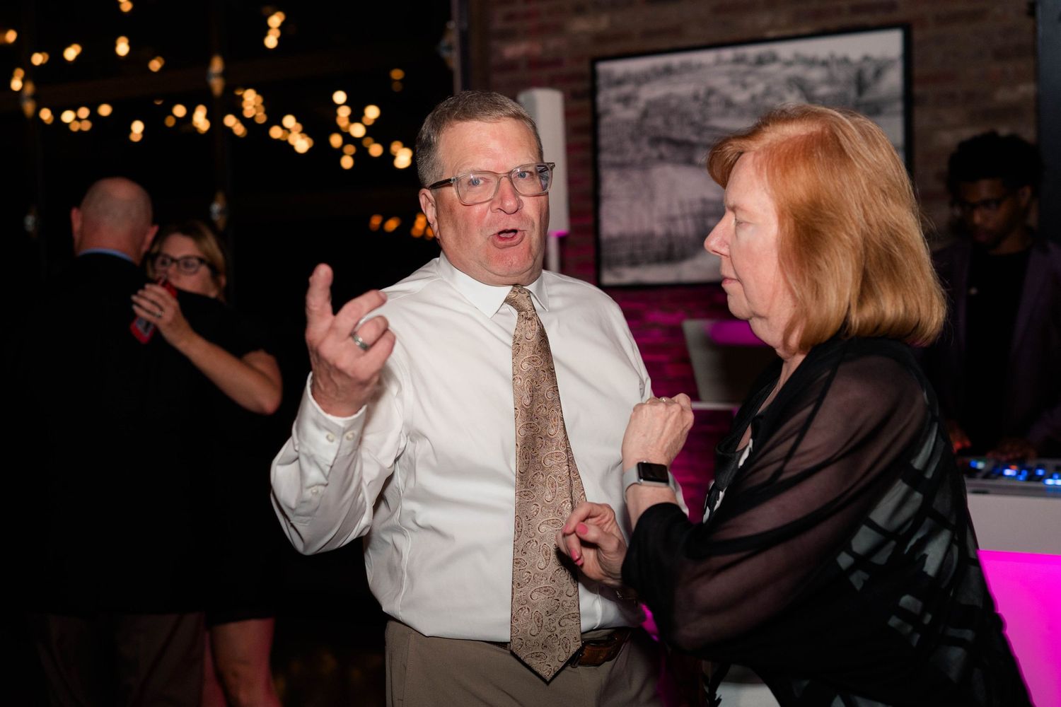 Older couple dancing and conversing at wedding reception with colorful DJ lighting.