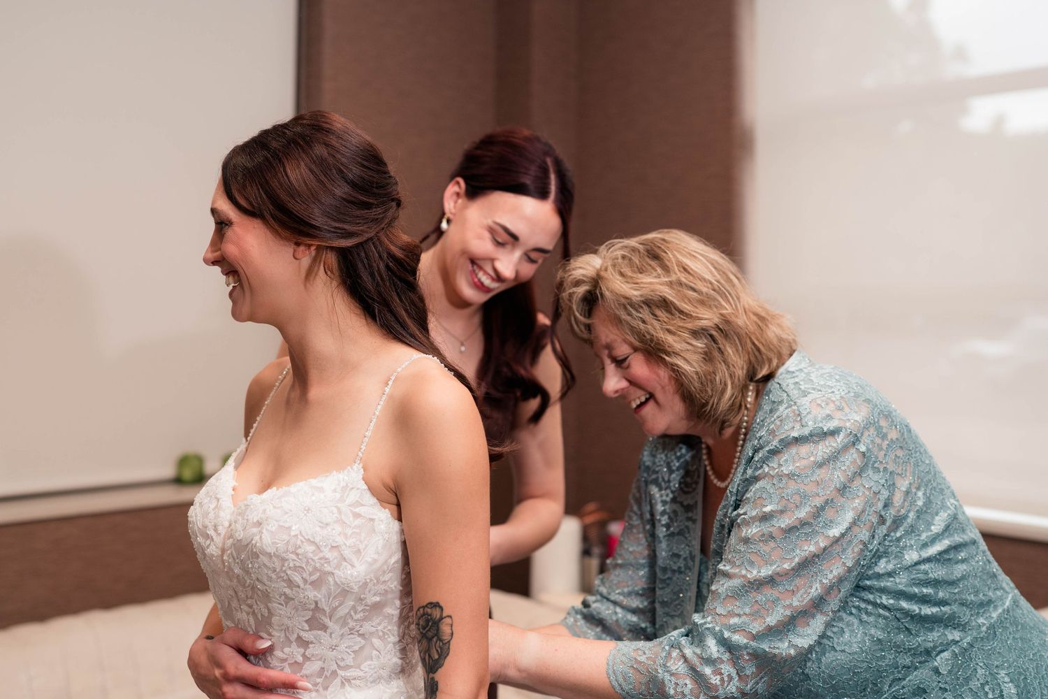 A bride in a white lace gown laughs as two women help her get dressed.