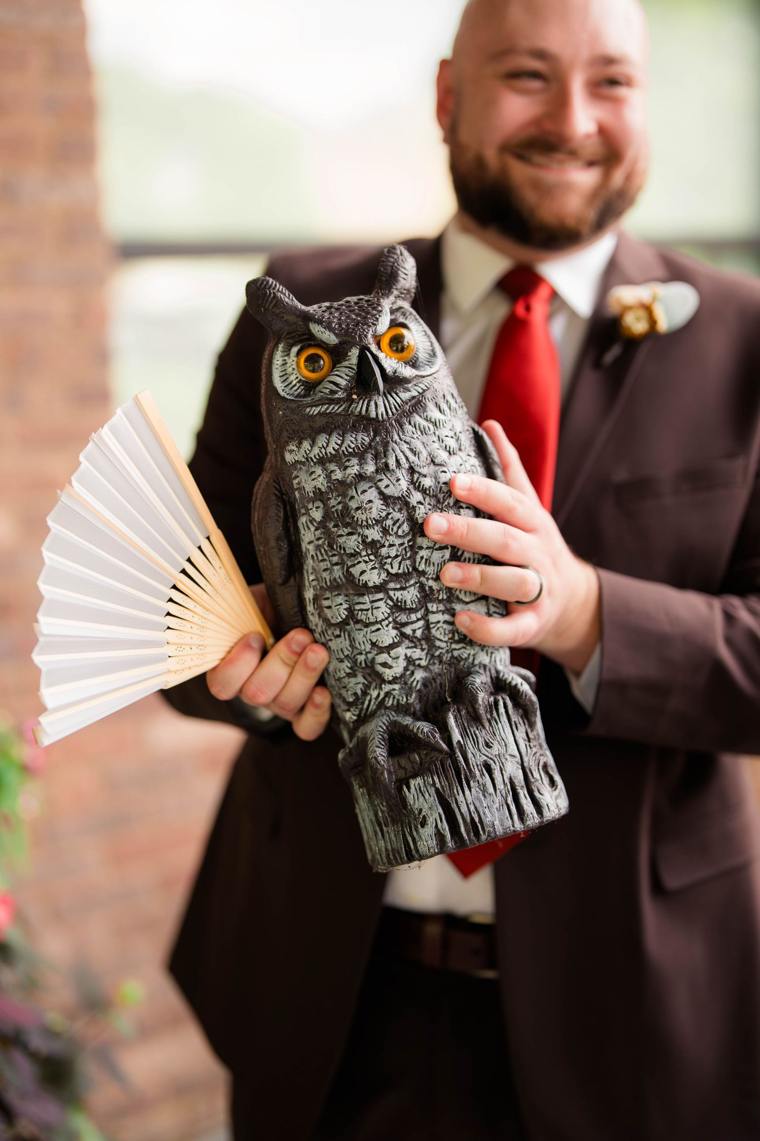 Smiling groom in brown suit and red tie holds decorative owl statue and paper fan at wedding.