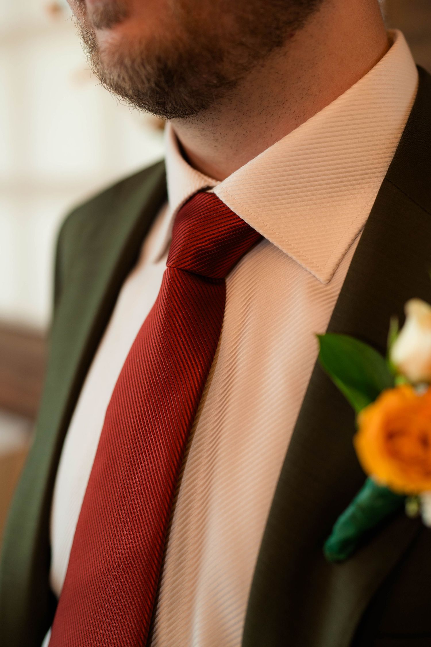 Close-up of groom wearing olive suit with burgundy tie and orange boutonniere on wedding day.