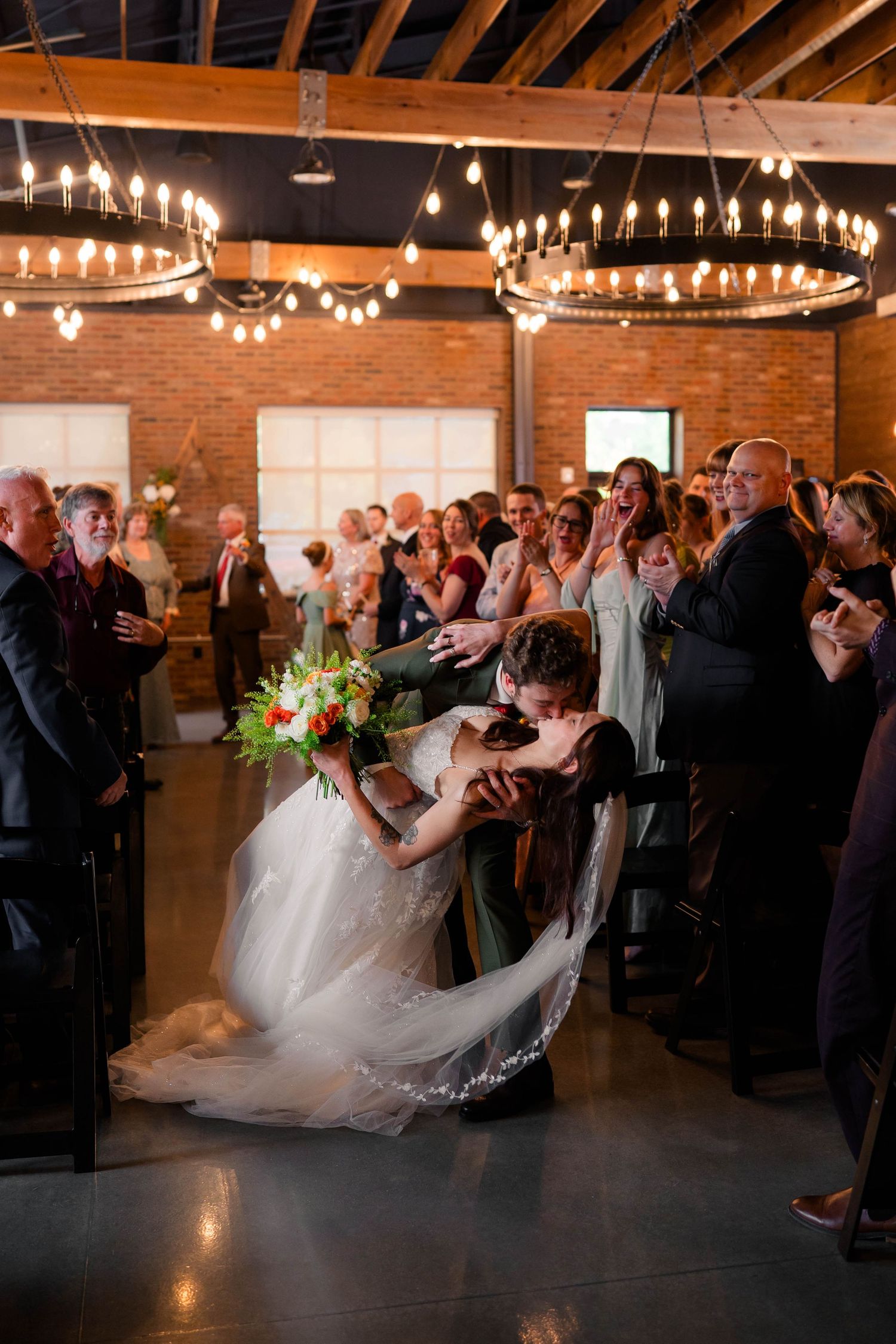 Groom dips bride for romantic kiss during recessional as wedding guests cheer in industrial venue.
