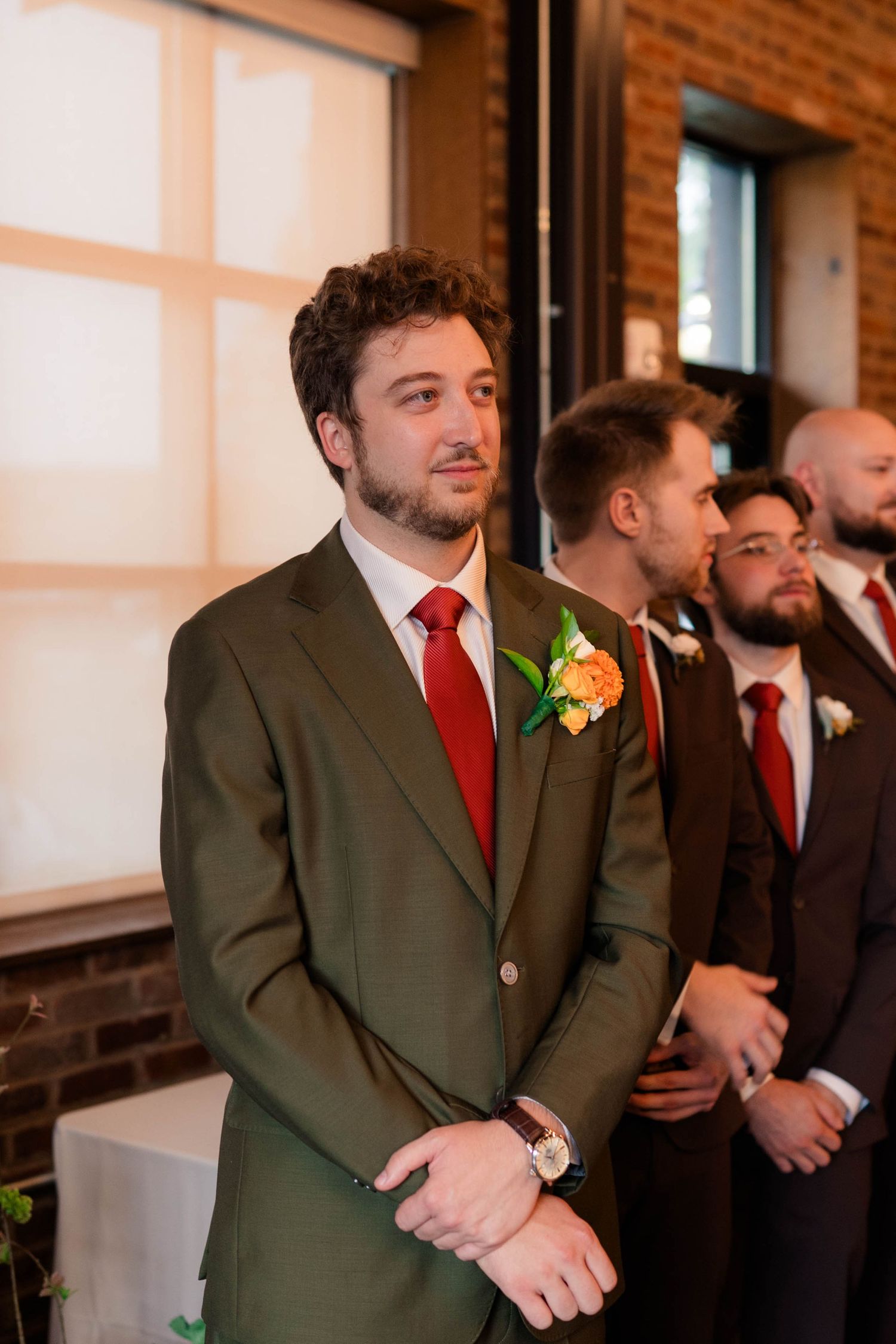 Groom in olive suit and red tie watches bride walk down the aisle with emotional expression.