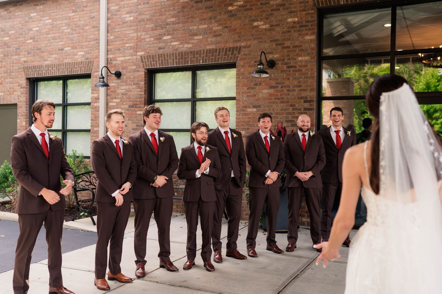 Groomsmen in dark suits with red ties reacting to bride during outdoor first look.