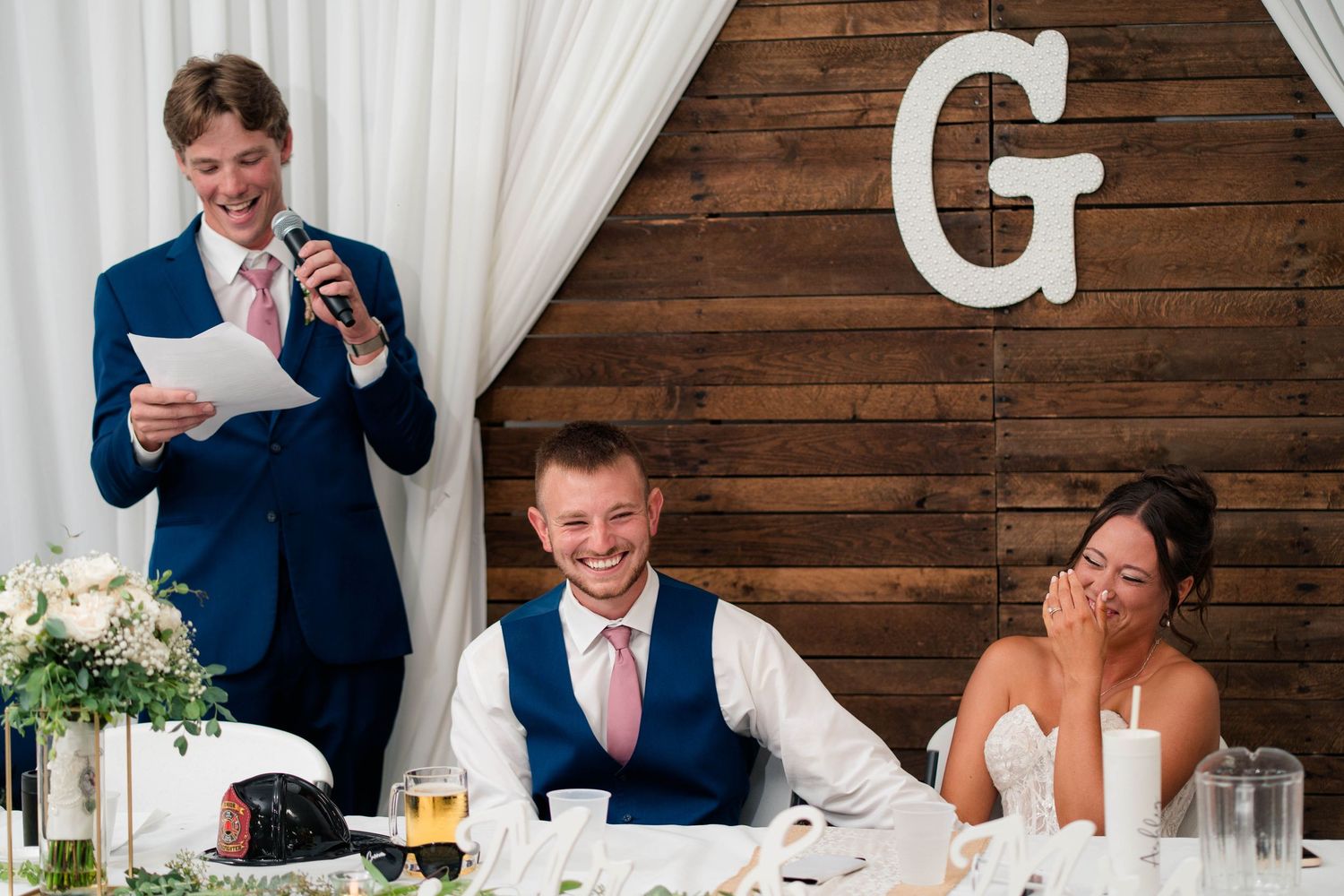 A groomsman delivers a humorous wedding speech as the smiling bride and groom sit at the head table beneath a wooden G monogram.