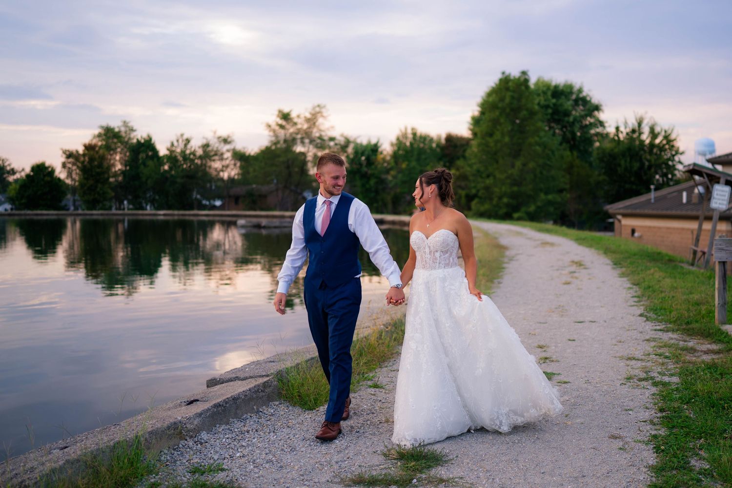 Happy newlyweds stroll together along a beautiful lakeside path during a golden sunset wedding photo.