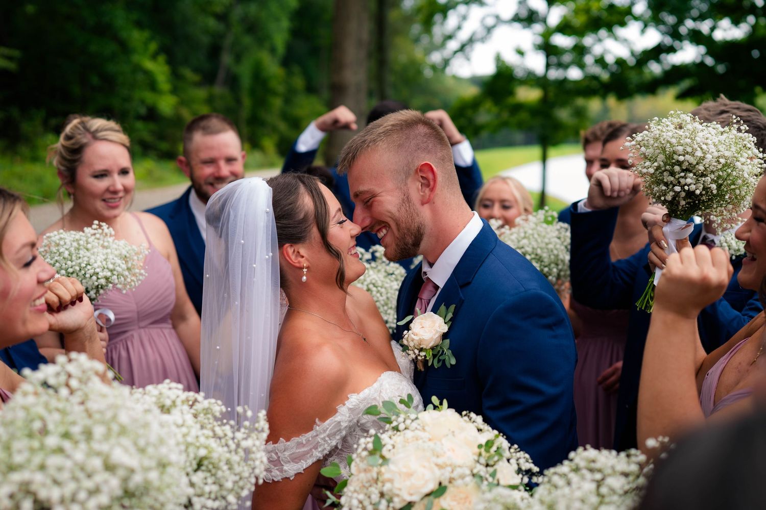 Bride and groom sharing a kiss surrounded by cheering wedding party holding baby's breath bouquets.