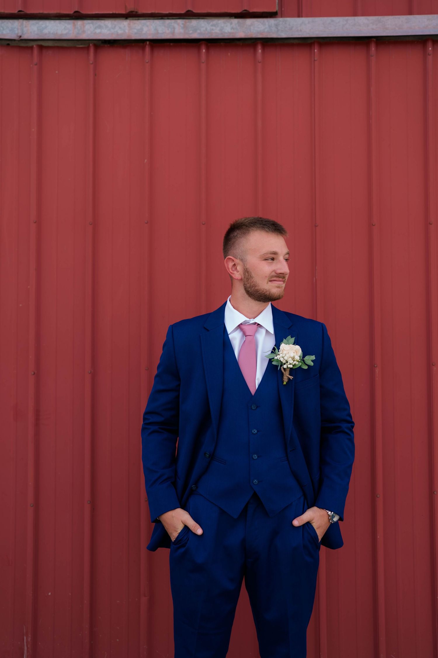 Handsome groom in navy blue three-piece suit with pink tie and boutonniere posing before red barn wall.