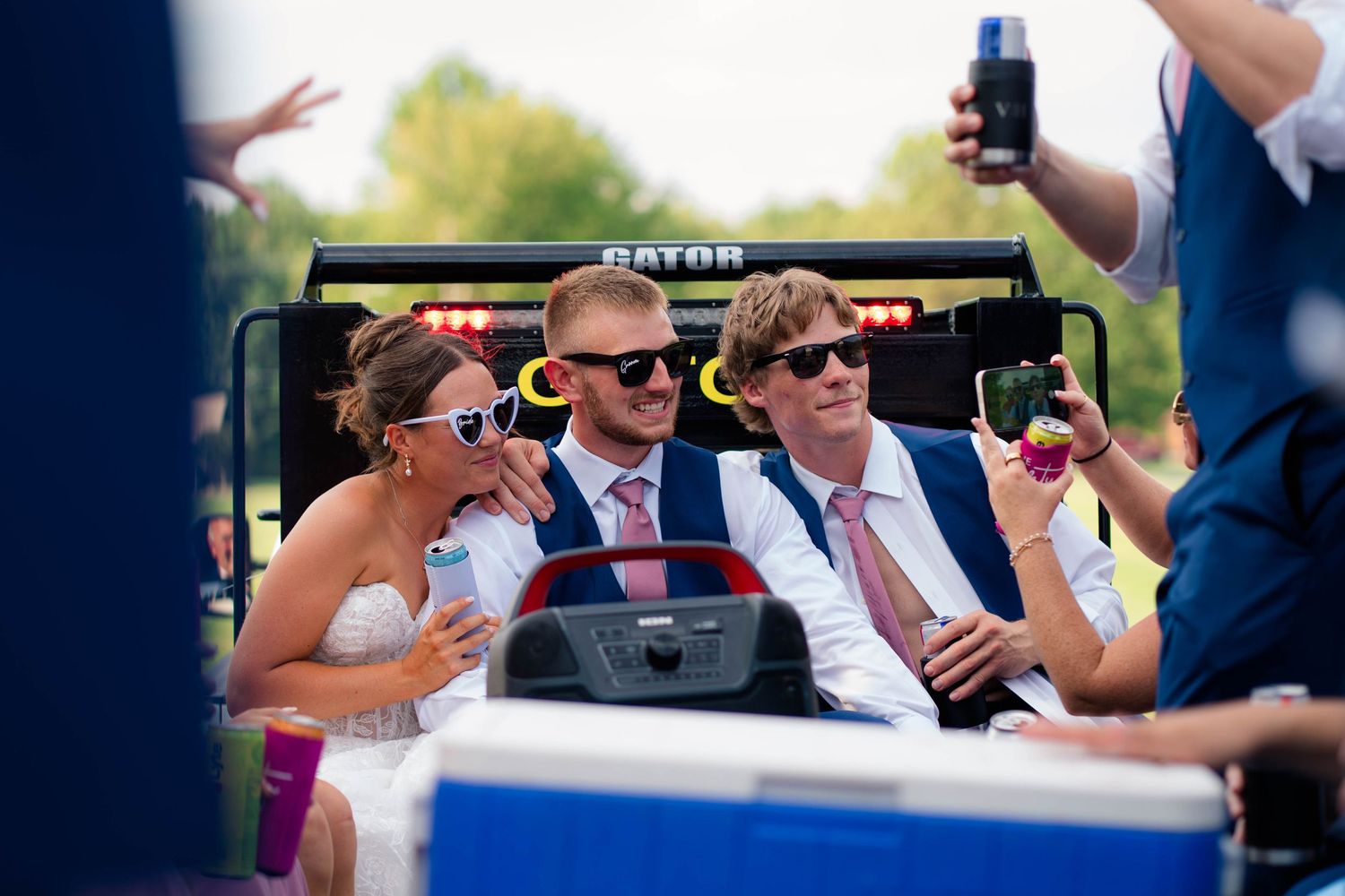 Bride and groom ride in Gator utility vehicle with guests wearing sunglasses at outdoor wedding celebration.