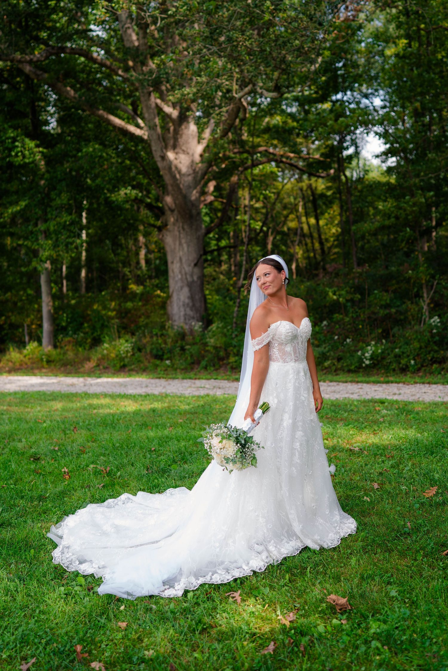 Elegant bride in off-shoulder lace wedding gown with cathedral train poses outdoors near large oak tree.