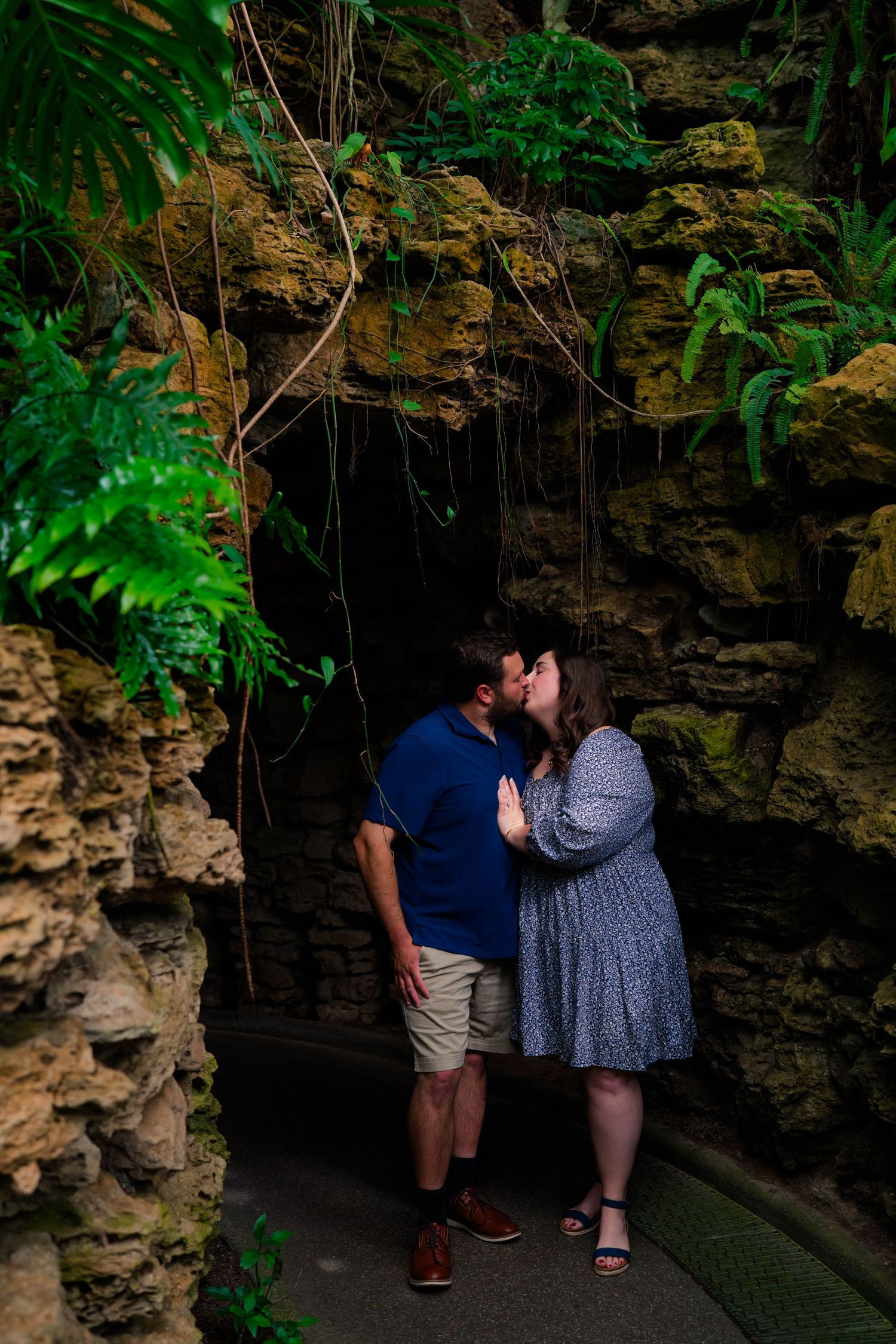 Romantic couple sharing a kiss in a cave-like setting surrounded by ferns and rocky walls.