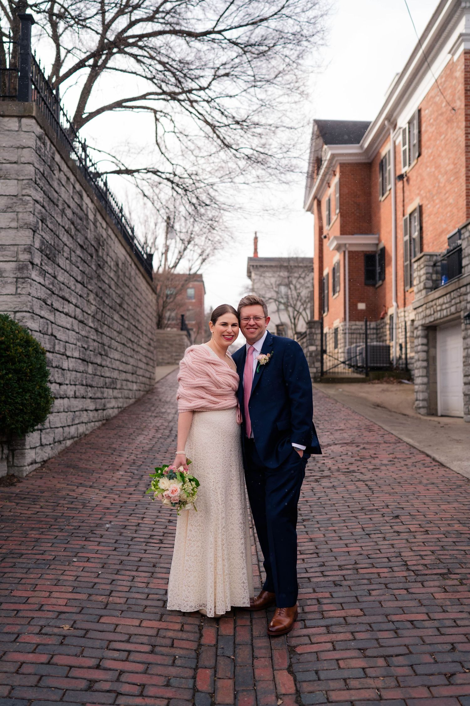 A couple in wedding attire poses on a brick alley, she in white with pink wrap, he in navy suit with pink tie.