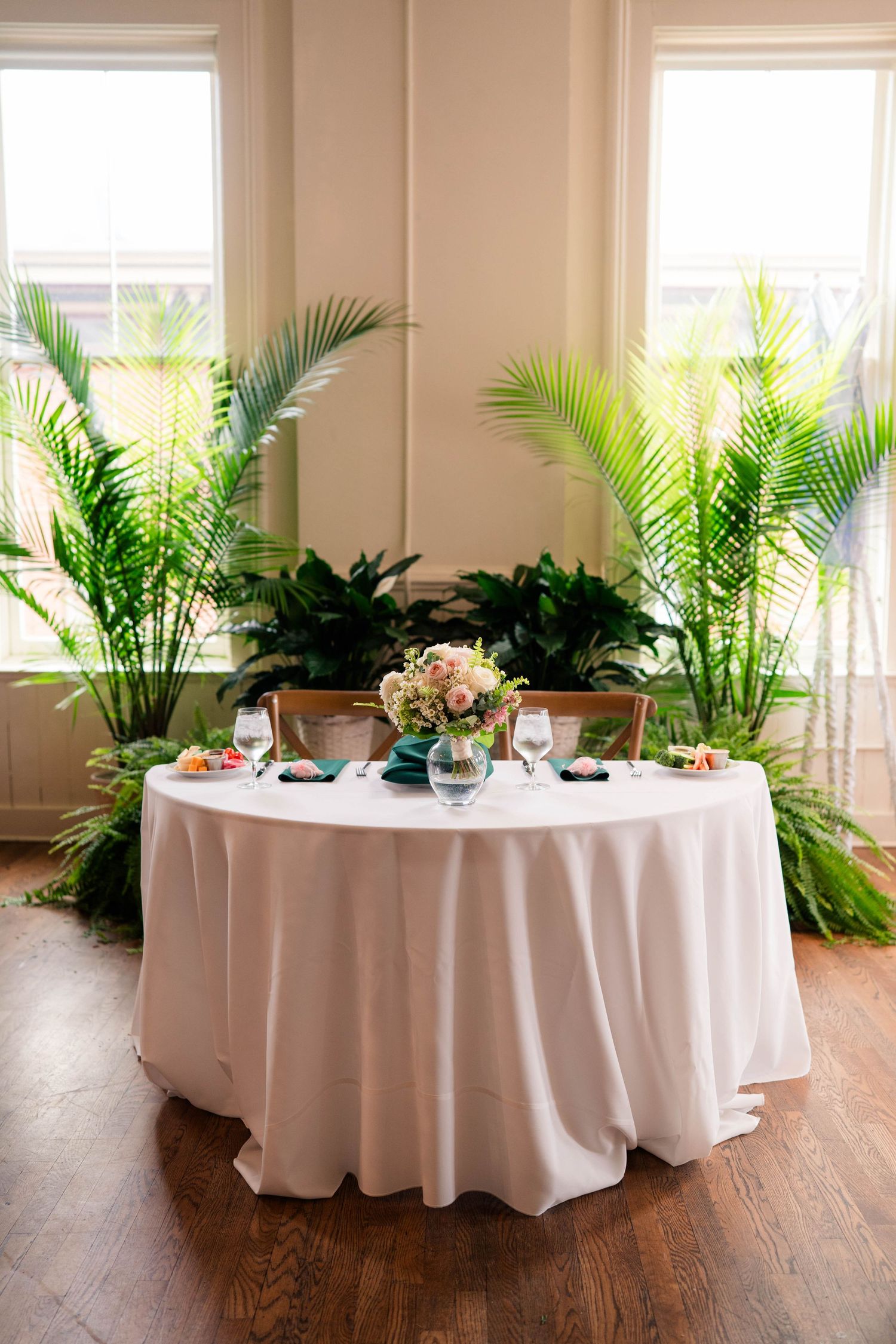 Elegant wedding sweetheart table with white linen, floral centerpiece, and lush tropical plants by sunlit windows.