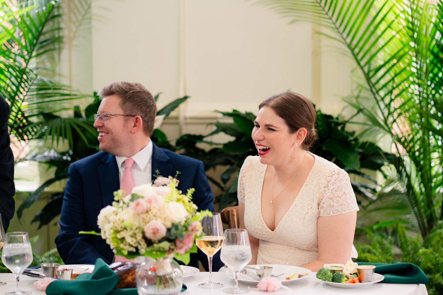 Bride and groom laughing at wedding reception dinner table with floral centerpiece and tropical greenery.