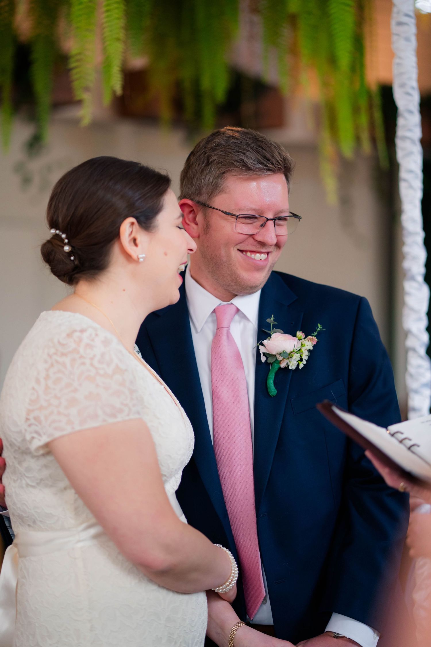 Bride and groom smile warmly at each other during wedding ceremony with officiant nearby.