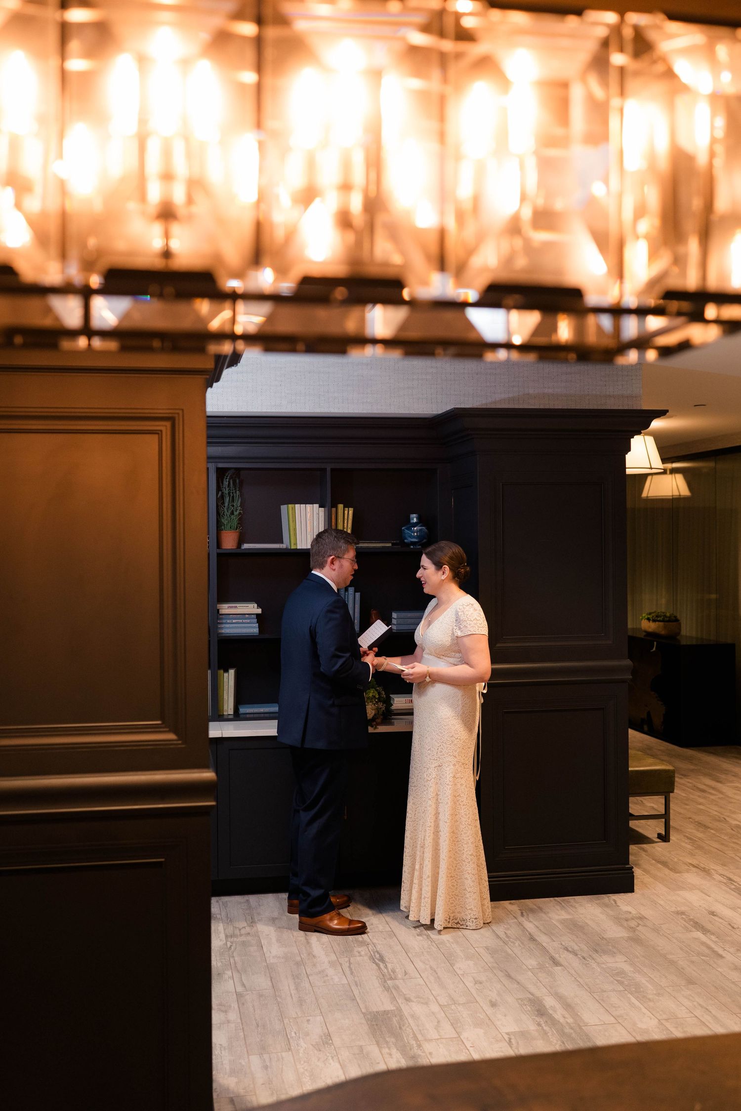 Couple exchanging private vows beneath warm glowing chandeliers in sophisticated hotel library setting.