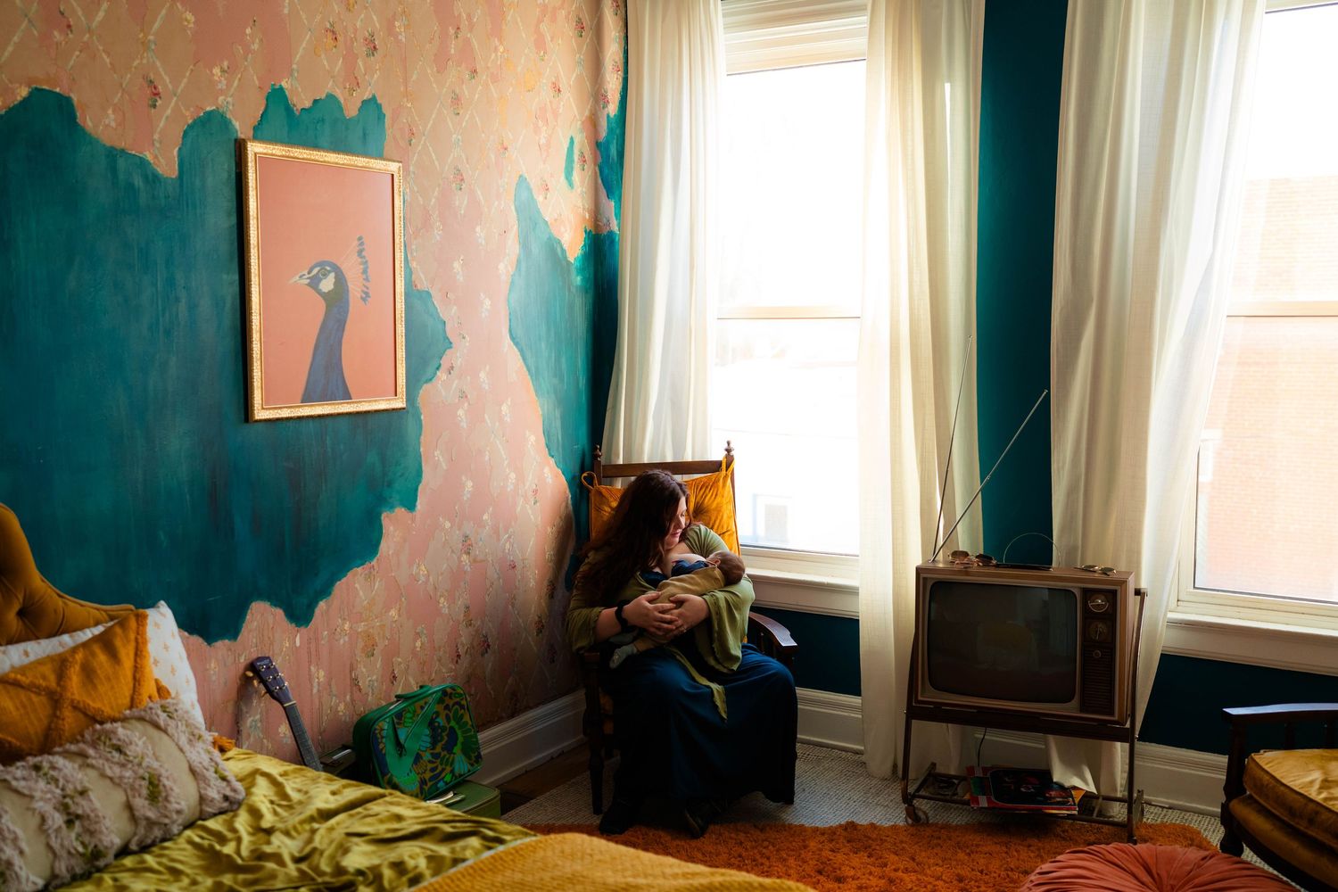 Mother nursing newborn baby in eclectic bohemian bedroom with teal painted walls by window.