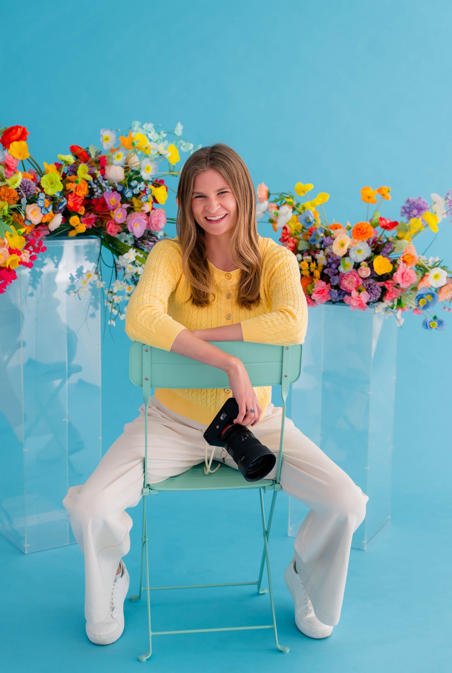 Smiling woman in yellow sweater sitting on mint chair holding camera with colorful flowers on blue background.