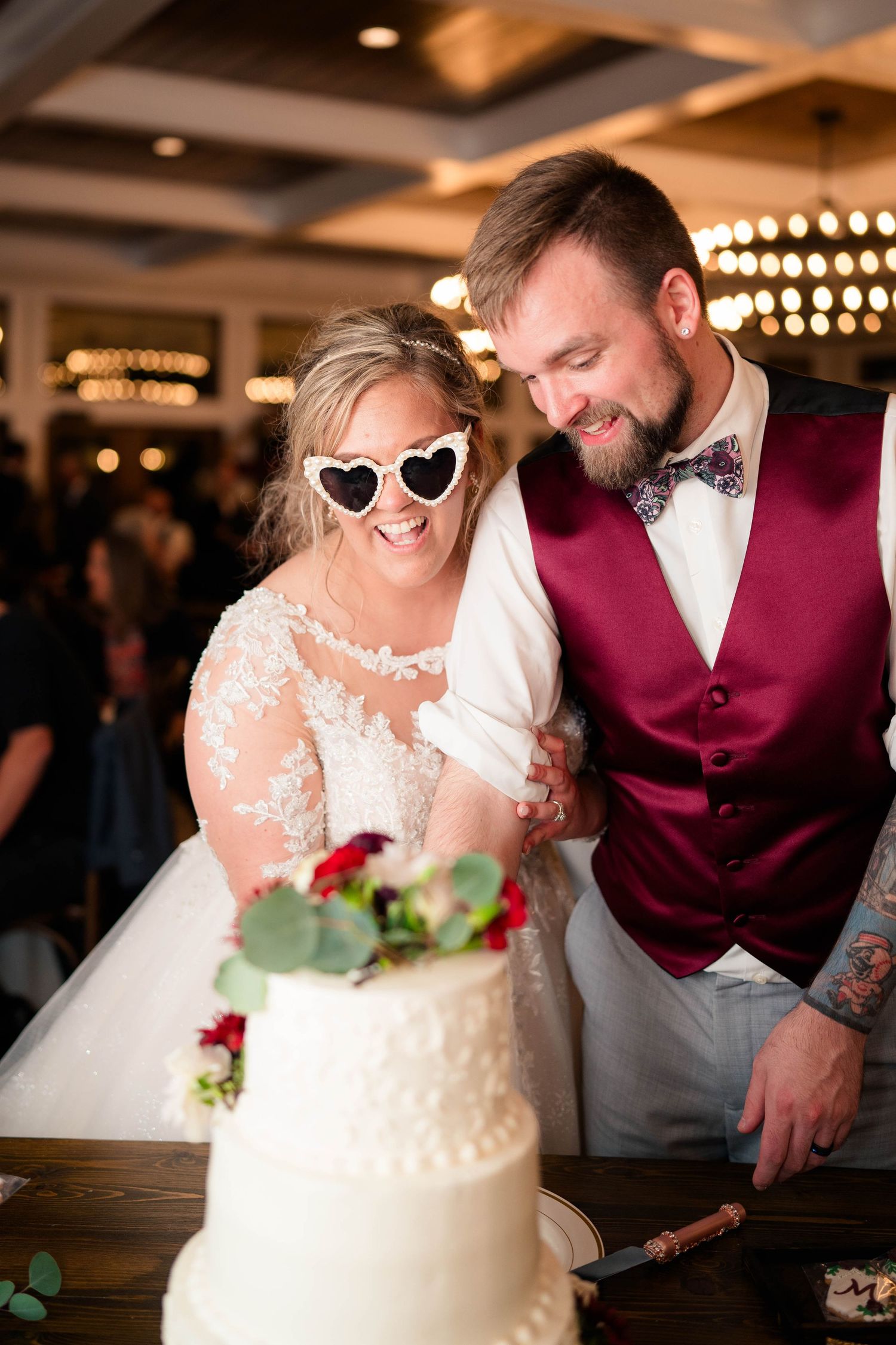 Bride wearing heart-shaped sunglasses and groom in burgundy vest cutting their white wedding cake together.
