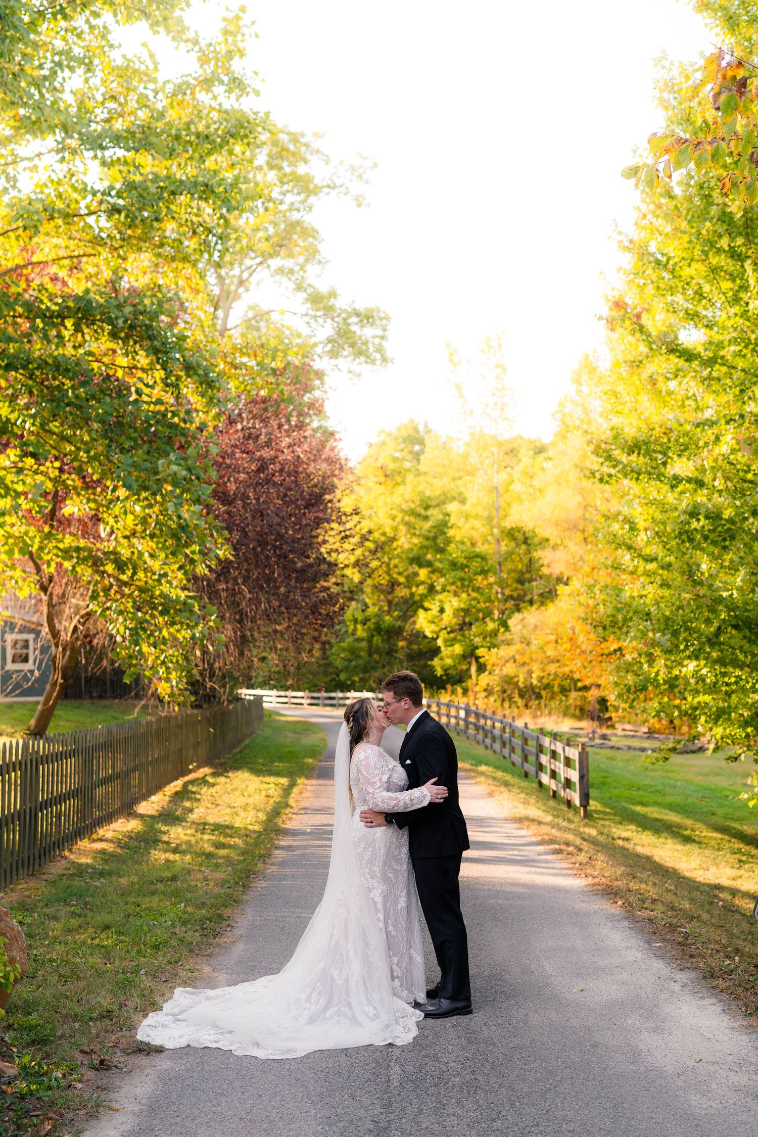Bride and groom share a romantic kiss on a sunlit tree-lined path during fall wedding portraits.