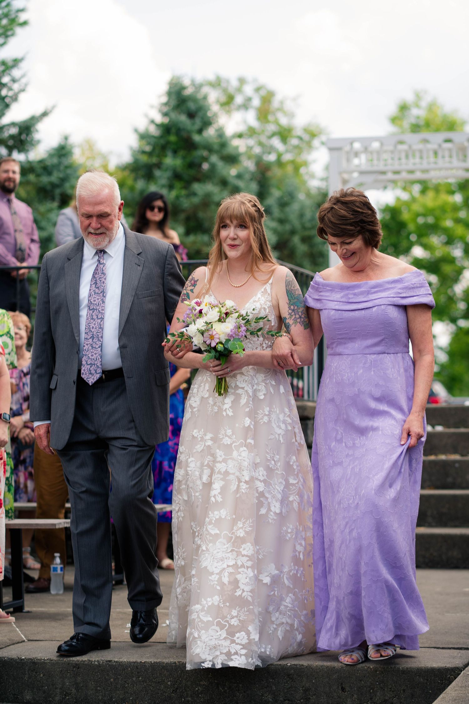 A bride in floral lace gown walks down the aisle escorted by her parents at an outdoor wedding.