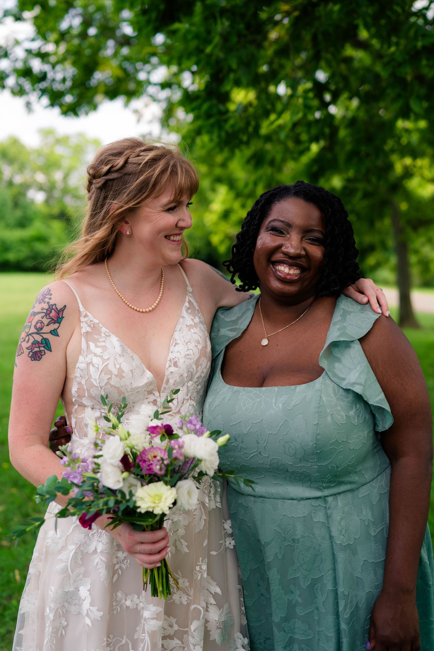 Bride with tattoo and bridesmaid in sage green dress smiling together outdoors.