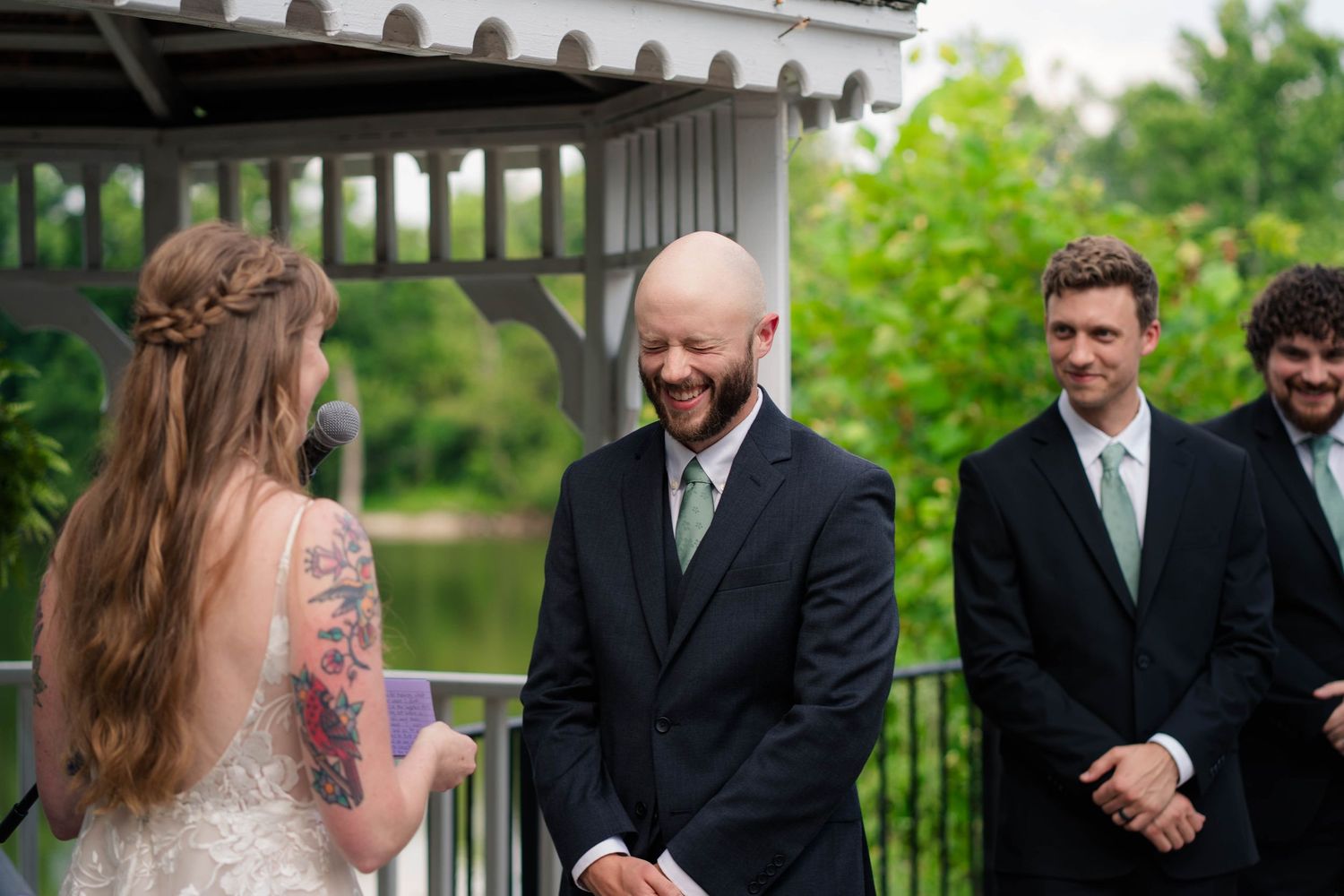 Bald groom smiles joyfully at bride during intimate outdoor gazebo wedding ceremony.
