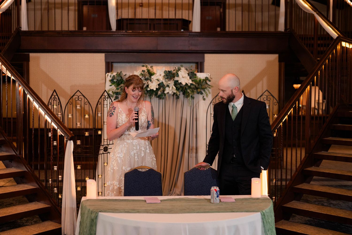 Bride and groom stand at sweetheart table beneath floral arrangement at indoor wedding reception.