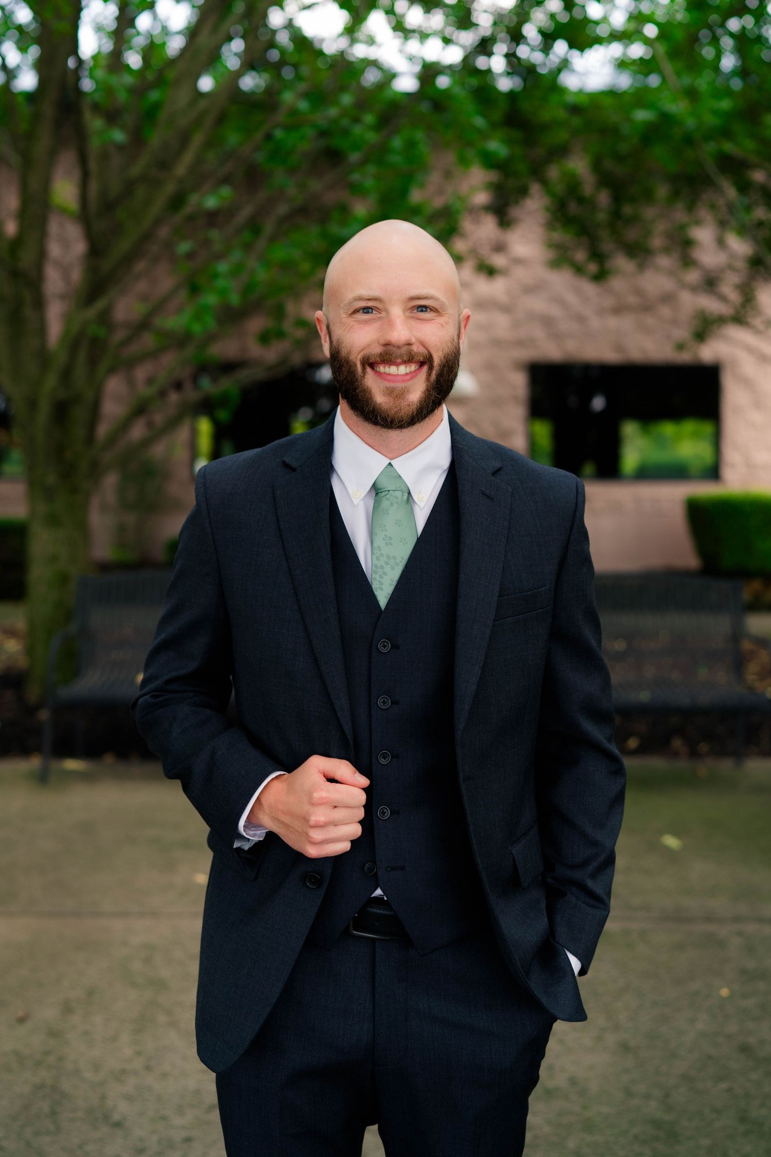 Confident bald groom with beard in navy three-piece suit and mint tie posing outdoors with hand in pocket.